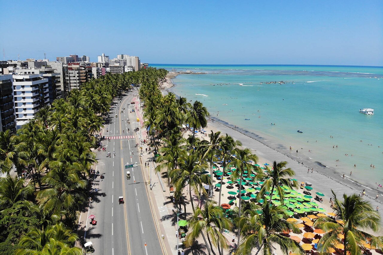 Vista aérea da orla de Ponta Verde, em Maceió, Alagoas, com avenida arborizada por coqueiros, prédios à esquerda e mar verde-azulado à direita